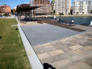 A waterfront promenade with paved walkways, a wooden deck, benches, a shaded structure, and buildings in the background.