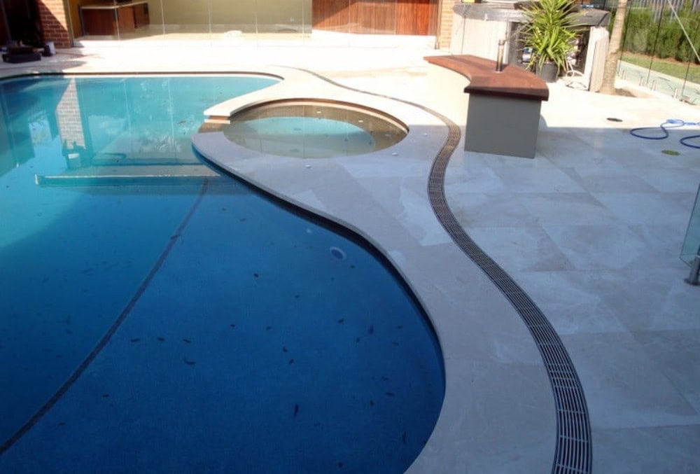 A modern backyard features a swimming pool with an attached circular spa, curved stone pool deck, and a glass fence in the background—perfect for relaxing, especially when regular strip drain maintenance keeps water flowing smoothly.