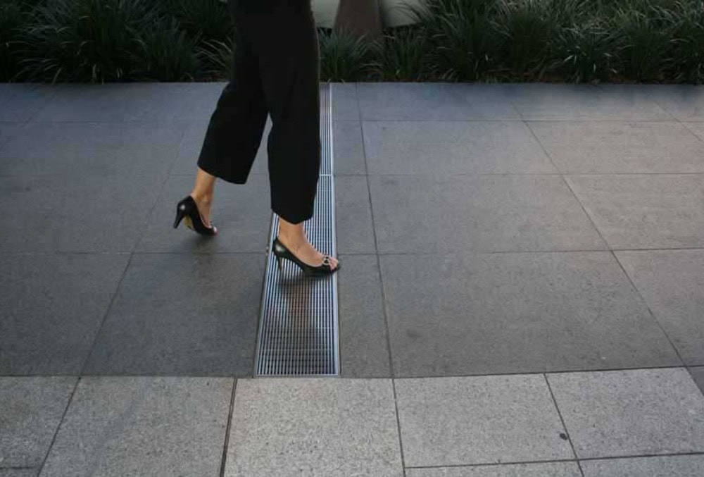 A person wearing black pants and black high-heeled shoes walks on a tiled sidewalk next to a metal grate—demonstrating how strip drains prevent surface pooling near the surrounding greenery.