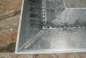 A close-up view of a metal floor grate set into a stone surface, showing water spraying over part of the grate—an ideal moment to consider linear drain maintenance or refer to your strip drain checklist.